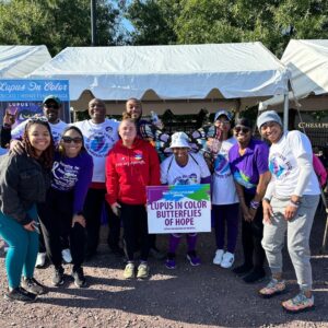 A diverse group of individuals stands together outdoors in front of white tents, participating in a lupus awareness event. They’re dressed in purple and white event shirts, with one person wearing a red hoodie that reads “you are enough.” The group is smiling and holding a sign that reads “Lupus in Color Butterflies of Hope.” Two people wear butterfly wings, and a banner for Lupus in Color is visible in the background. The setting is sunny, with trees providing a green backdrop.