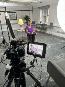 A behind-the-scenes photo of a professional video interview setup. A woman wearing a purple shirt sits on a gray chair in a well-lit room, facing a camera. She appears on the monitor of the camera setup in the foreground. Lighting equipment, a boom microphone, and a monitor are clearly visible, capturing the scene. The setting includes a lamp, framed artwork, and neatly arranged shelves and chairs in a contemporary space with natural light from a large window.